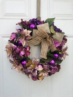 a wreath with purple flowers and green leaves hanging on the front door to welcome guests