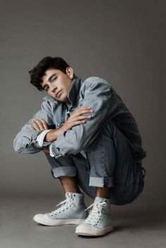 a young man sitting on the ground with his arms crossed, wearing white sneakers and denim jacket