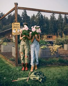 two people standing in front of a fence with flowers on their heads and holding plants