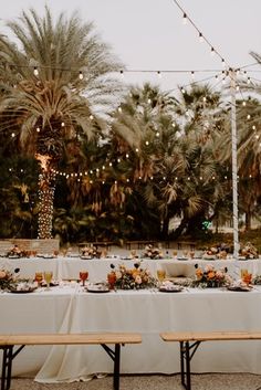 a long table is set up with white linens and lights strung from the palm trees