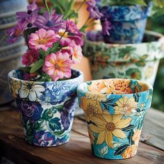 four colorful flower pots sitting on top of a wooden table with flowers in the background