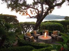 an outdoor fire pit surrounded by bushes and trees with the ocean in the back ground