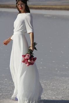 a woman in a long white dress holding a bouquet of flowers and walking on the beach