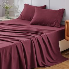 a bed covered in maroon sheets and pillows next to a night stand with flowers on it