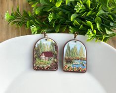 pair of earrings with scenic scene painted on them sitting on a white plate next to green plants