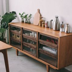 a wooden sideboard with drawers and plants on top in a white walled room next to a table