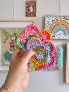 a person holding up a crocheted flower in front of pictures on the wall