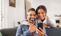 a man and woman sitting on a couch looking at a cell phone while they smile