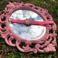 a pink mirror sitting on the ground with a watch in it's reflection,