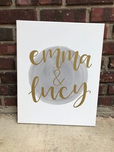 a white canvas with gold foil lettering on it that says emma and lucy next to a brick wall