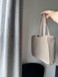 a hand holding a crocheted bag in front of a window with curtains behind it