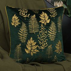 a green pillow with gold leaf designs on it sitting on a chair in front of a potted plant