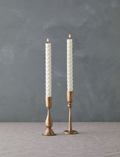 two white candles sitting next to each other on top of a wooden table in front of a gray wall