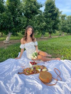 a woman sitting on a blanket in front of an apple orchard with fruits and cheeses