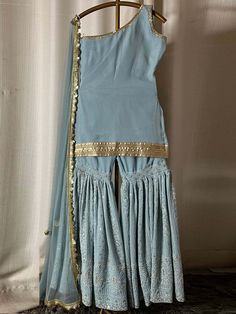 This is a three-piece grey blue sharara set from the Glitter Choli collection. This gorgeous grey blue georgette one-shoulder kurta is paired with an embroidered georgette sharara pants. The grey blue sharara set is enhanced with heavy sequin embroidery and gold sari lace work. The mid-thigh fitted sharara pants have pleats with heavy embroidery. The outfit is completed with a sequin work grey blue dupatta in net material with scalloped edging. Unstitched Light Blue Georgette Sets, Light Blue Georgette Bollywood Sharara, Light Blue Bollywood Style Georgette Sharara, Light Blue Georgette Sets For Eid, Light Blue Floor-length Sharara For Festive Occasions, Fitted Light Blue Sharara For Eid, Light Blue Georgette Party Set, Light Blue Resham Embroidered Sharara For Party, Anarkali Light Blue Sharara For Party