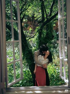 a man and woman are kissing in front of an open window with trees behind them