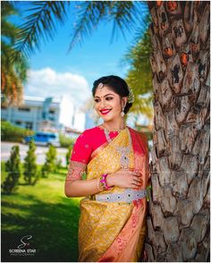 a woman standing next to a palm tree wearing a yellow and pink sari with red accents