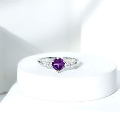 a purple heart shaped ring sitting on top of a white stand with three diamonds around it