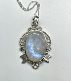 This enchanting pendant showcases a mesmerizing rainbow moonstone with an iridescent glow, set in a beautifully detailed sterling silver setting adorned with celestial stars and a crescent moon. Suspended on a 20" sterling silver chain, this piece captures the magic of the night sky, making it a perfect gift for dreamers and stargazers. The Moonstone approximately 1 7/8" long and 9/16" wide. Known for its soothing energy and ability to restore balance, the rainbow moonstone is not only lovely to Mystical Oval Moon Phase Jewelry, Celestial Silver Moon Crystal Necklace, Celestial Moon Phase Crystal Necklaces, Healing Moonstone Crystal Necklace With Moon Phase, Healing Moonstone Moon Phase Crystal Necklace, Mystical Iridescent Moonstone Jewelry, Celestial Moon Phase Necklace In Labradorite, Celestial Silver Gemstone Crystal Necklaces, Celestial Moon Phase Crystal Necklace In Silver