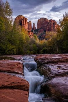 a river running through a lush green forest filled with red rocks and tall mountains in the background