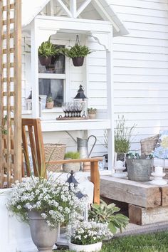 a white house with potted plants and flowers on the front porch, next to a wooden bench