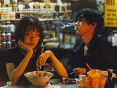 two people sitting at a table with food in front of them and one person holding chopsticks to their mouth