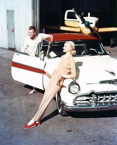 an old photo of a woman leaning on the hood of a car with another man standing next to her