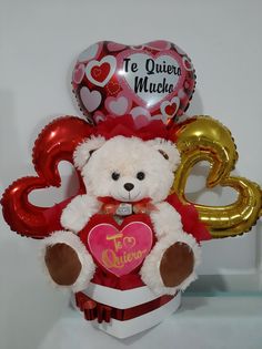 a white teddy bear sitting on top of a heart shaped box filled with foil balloons