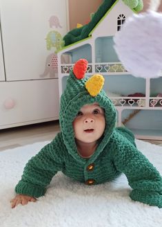 a baby wearing a green crochet costume laying on the floor in front of a toy house