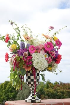 a vase filled with lots of colorful flowers on top of a brick wall next to trees