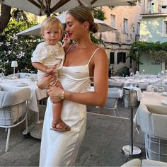 a woman holding a small child in her arms at an outdoor dining area with tables and chairs