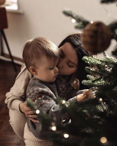 a woman is holding a baby near a christmas tree with lights on it and kissing her cheek