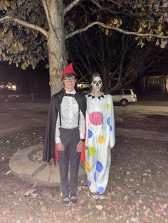 two people dressed up as clowns standing next to each other in front of a tree