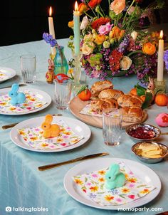 the table is set for easter dinner with colorful flowers and bunnies on plates, candles, and croissants