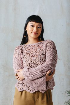 a woman standing in front of a wall wearing a pink crochet sweater and brown pants