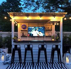 an outdoor bar with stools and lights on the outside, in front of a fence