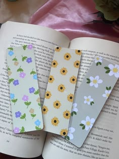 three bookmarks sitting on top of an open book next to flowers and a candle