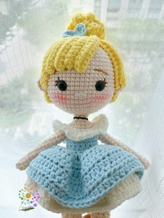 a crocheted doll with a blue dress and yellow hair is posed in front of a window