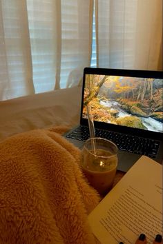 an open laptop computer sitting on top of a bed next to a book and drink