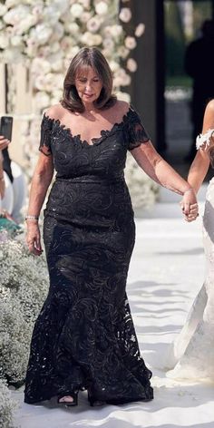 two women in black dresses walking down the aisle with one woman holding her hand and smiling at the camera