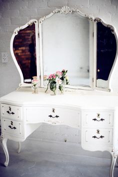 an antique white dressing table with mirror and flowers on the top, in front of a brick wall