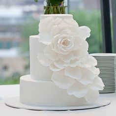 a three tiered white wedding cake with flowers on the top and bottom, in front of a window