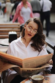 a woman reading a newspaper while sitting at a table with a cup of coffee in front of her