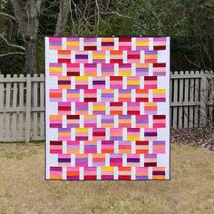 a colorful quilt on the ground in front of a white fence and trees with grass