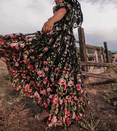 a woman in a black floral print dress is standing by a fence and looking at the camera