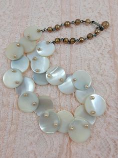 "This is a vintage, perhaps handmade, necklace with a brass chain and hook. The necklace is comprised of Mother of Pearl shell discs, strung through their 2 holes to connect with one another. The front shell discs are topped with ivory colored faux pearl beads. The ends of the necklace have short lengths of brass chain, interspersed with painted faux pearl beads in a darker color. The necklace has a hook style connector, and the length is adjustable. The necklace measures 16\" in length, so pret White Shell-shaped Mother Of Pearl Beaded Necklaces, Mother Of Pearl Shell-shaped Necklace In Pearl White, Vintage Mother Of Pearl Shell Jewelry, Adjustable Shell-shaped Mother Of Pearl Necklace, Adjustable Shell-shaped Mother Of Pearl Jewelry, Flapper Necklace, Pearl Shell, Gold Ribbons, Ivory Color