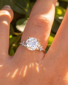 a woman's hand with a diamond ring on top of her finger and leaves in the background