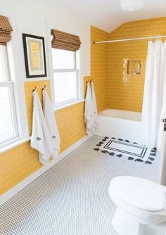 a white toilet sitting next to a bath tub under two windows in a bathroom with yellow walls