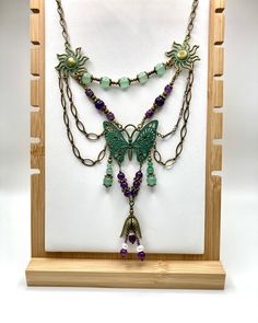 This stunning chandelier necklace features an intricate metal butterfly at its center, adorned with a beautiful patina wash in green. The butterfly is connected to strands of green aventurine and amethyst, which gracefully lead up to the sun charms on either side. The necklace is tied together by a gorgeous horse-eye chain in antique bronze, draped twice on both sides. At the bottom of the necklace, there is a striking flower bead that serves as the lower focal point. The necklace measures 17 inches in length and comes with a 1-inch extender chain, finished with an adorable metal bee bead that adds a touch of whimsy. Bohemian Necklace With Butterfly Charm, Bohemian Dangle Jewelry With Butterfly Charm, Bohemian Jewelry With Butterfly Charm Dangle, Bohemian Butterfly Jewelry, Bohemian Butterfly Charm Jewelry For Jewelry Making, Bohemian Jewelry With Butterfly Charm For Jewelry Making, Gold Bohemian Butterfly Necklace, Bohemian Butterfly Necklace For Gift, Handmade Bohemian Butterfly Necklace