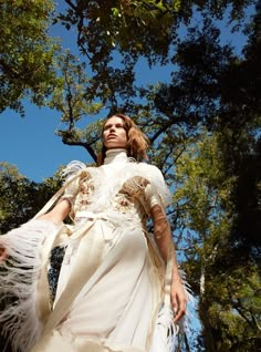 a woman in a white dress standing next to trees and looking up at the sky